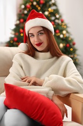Photo of Beautiful young woman in Santa hat on sofa near Christmas tree at home