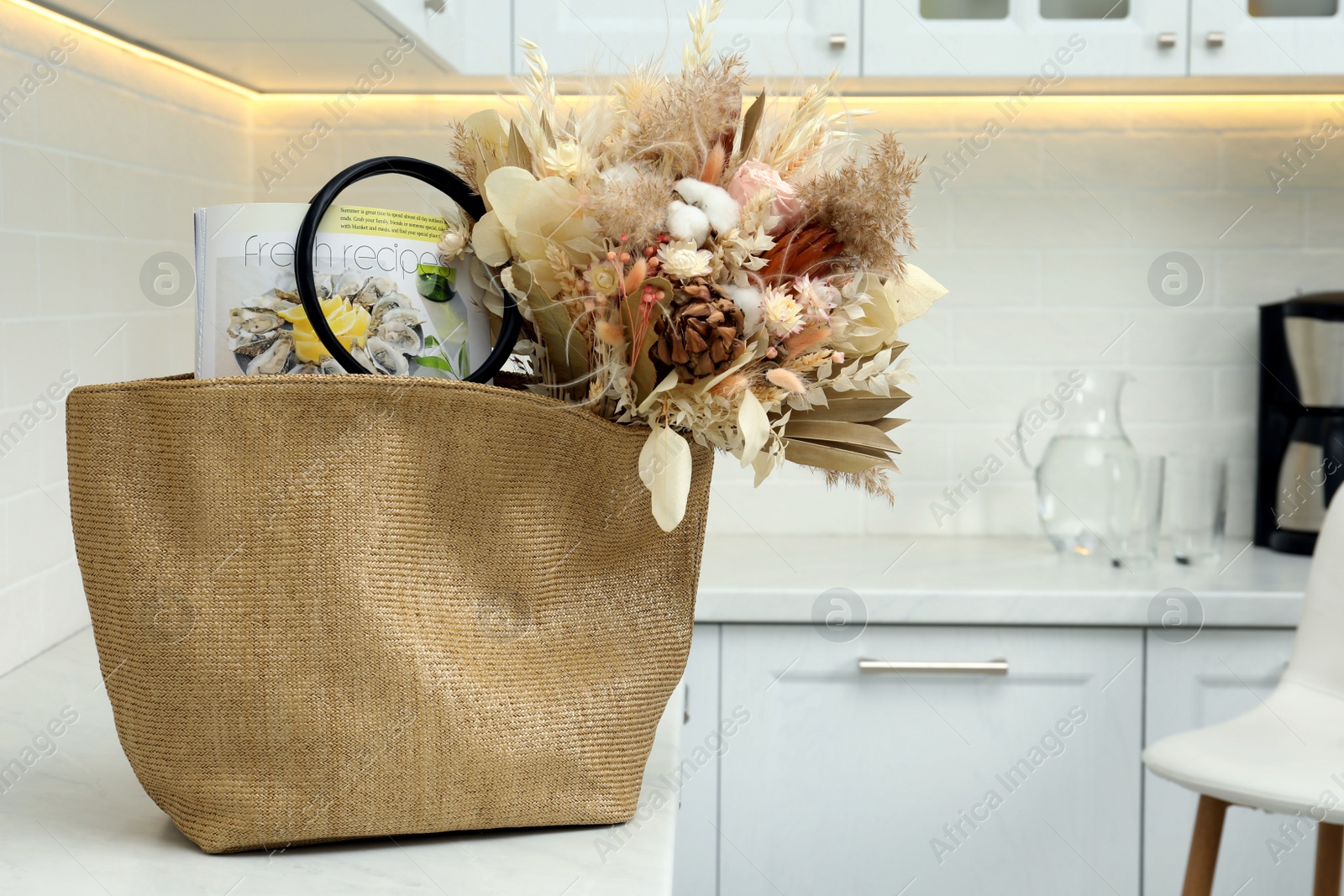 Photo of Stylish beach bag with beautiful bouquet of dried flowers and magazine on countertop in kitchen. Space for text