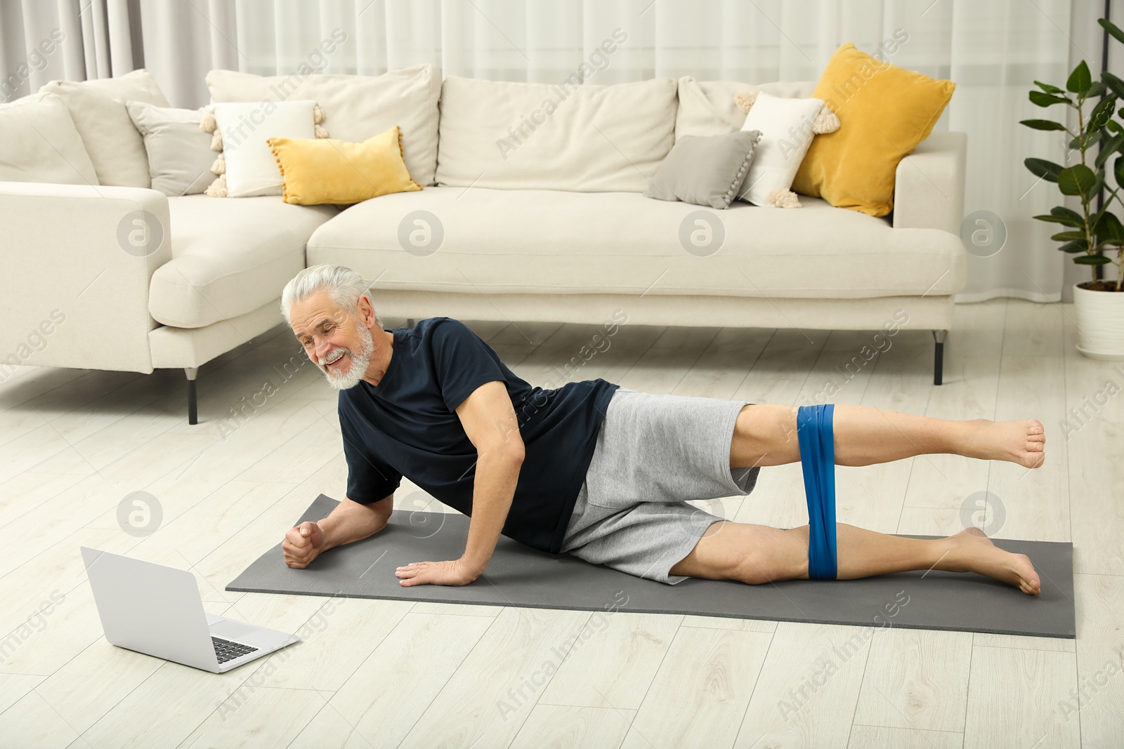 Photo of Senior man doing exercise with fitness elastic band near laptop on mat at home