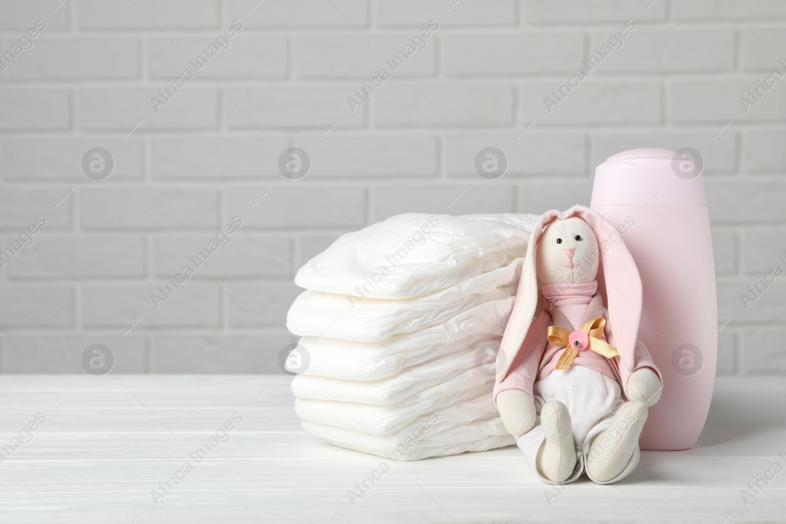 Photo of Baby diapers, toy bunny and bottle on wooden table against white brick wall. Space for text