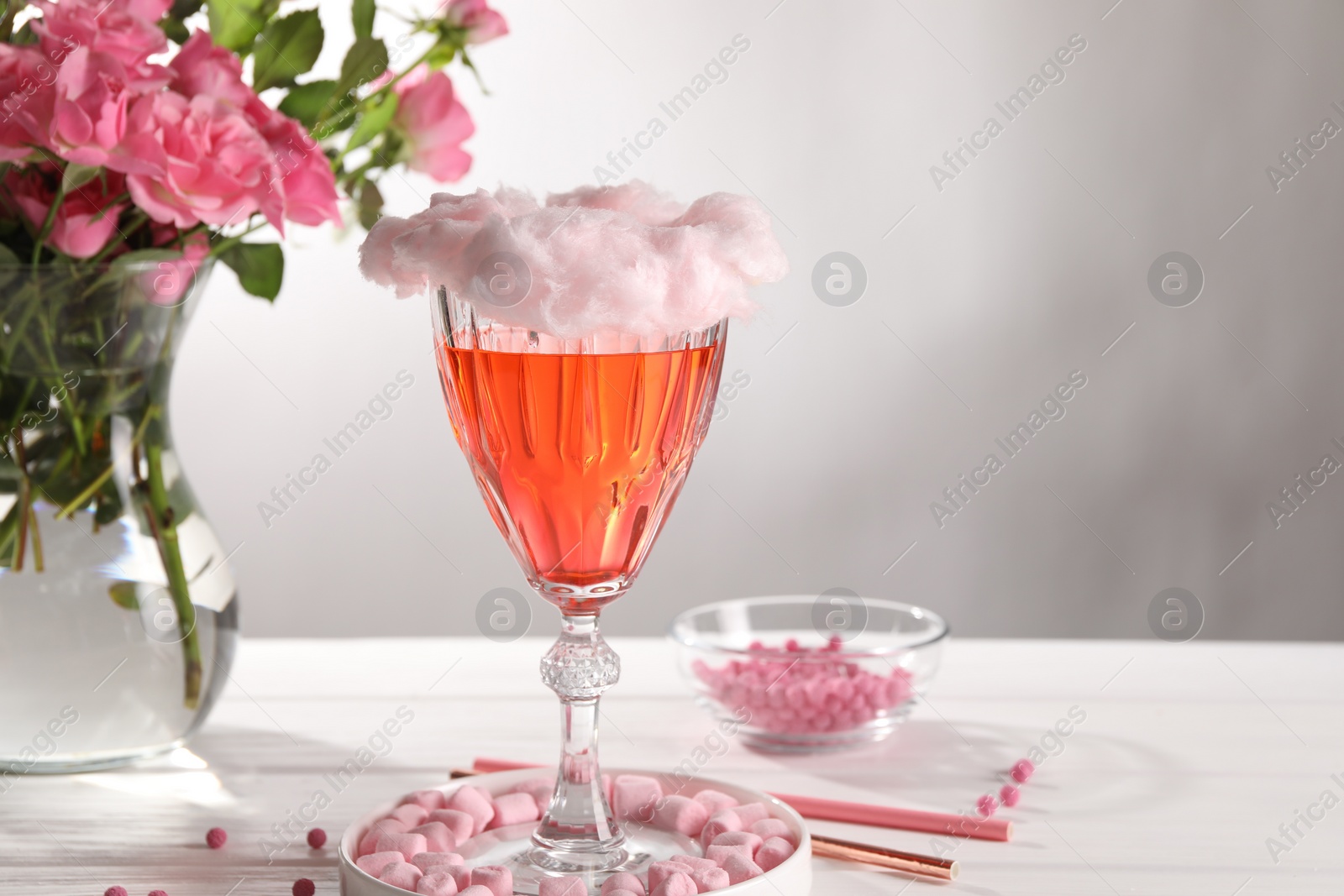 Photo of Cotton candy cocktail in glass, marshmallows, vase with pink roses and straws on white wooden table against gray background, space for text