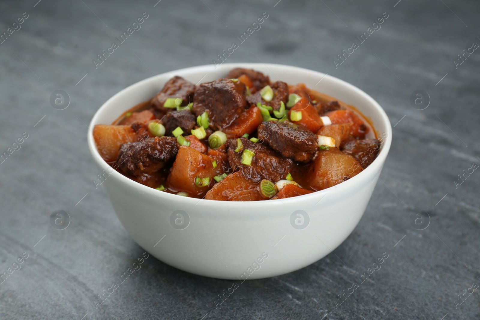 Photo of Delicious beef stew with carrots, green onions and potatoes on grey table, closeup