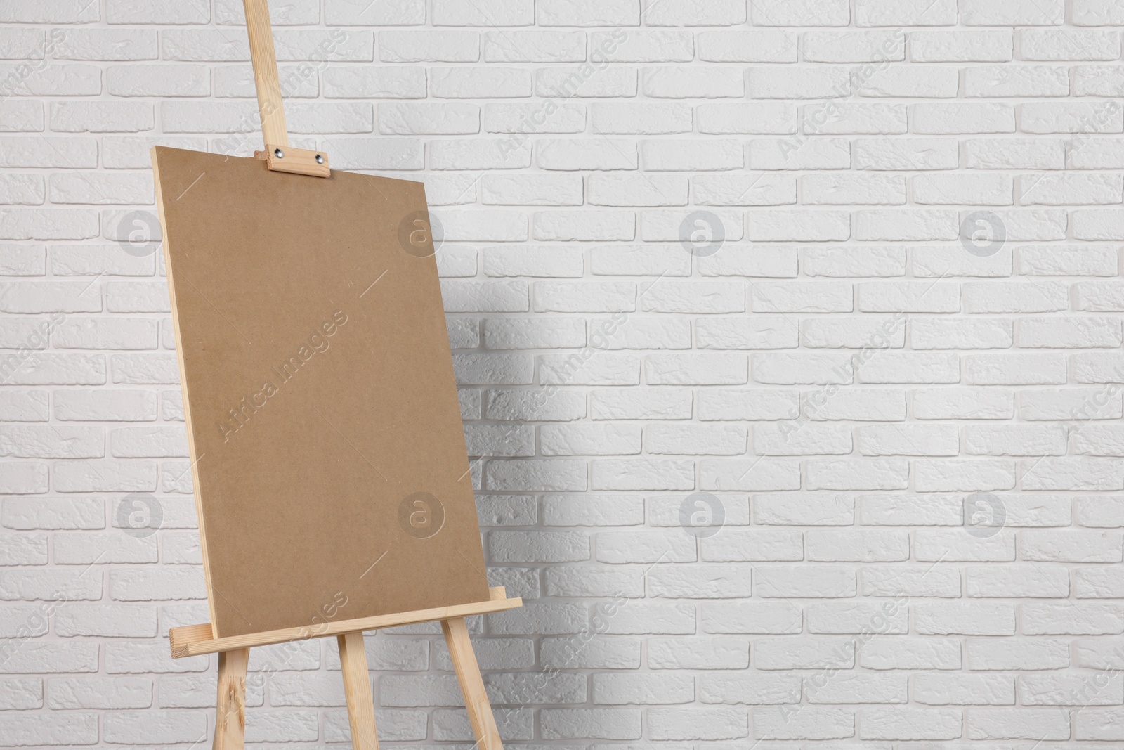 Photo of Wooden easel with blank board near white brick wall. Space for text