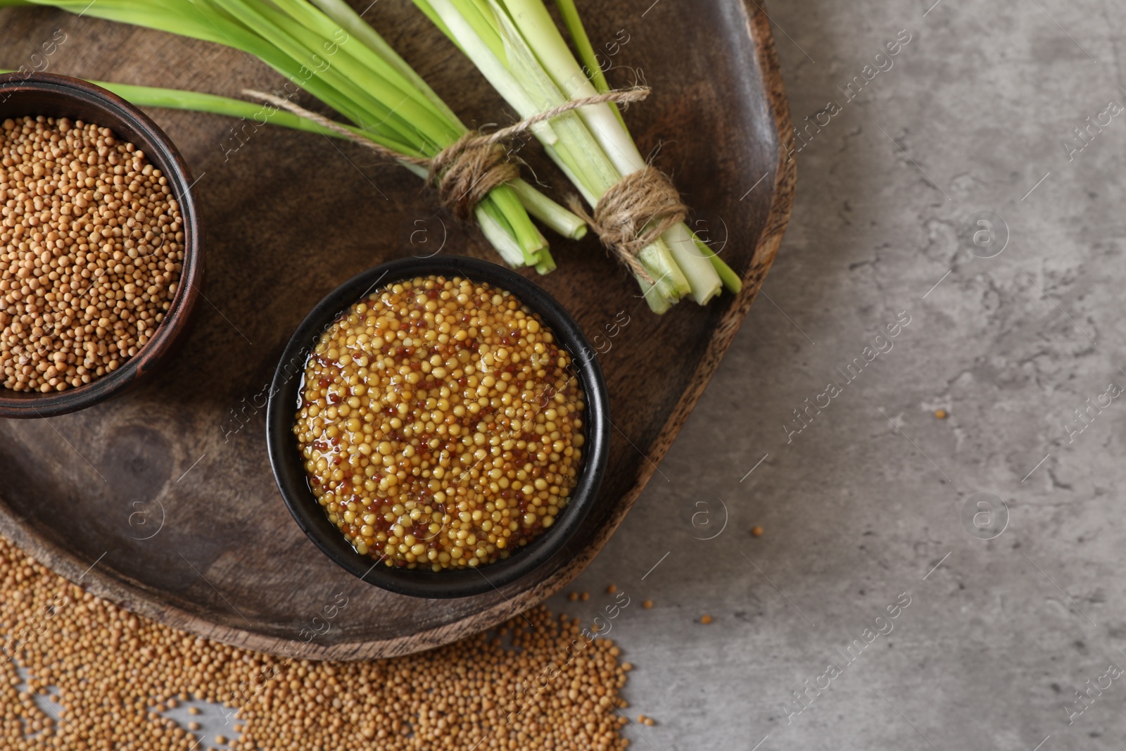 Photo of Serving board with delicious whole grain mustard, seeds and fresh green onion on grey table, top view. Space for text