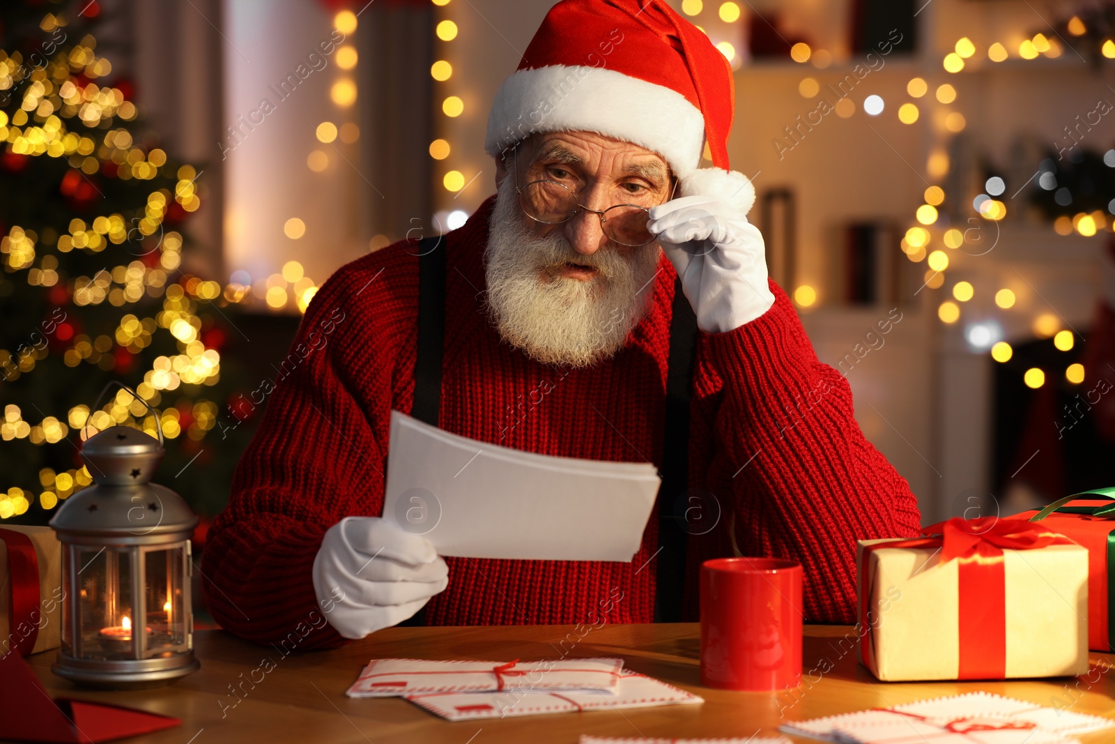 Photo of Santa Claus reading letter at his workplace in room with Christmas tree