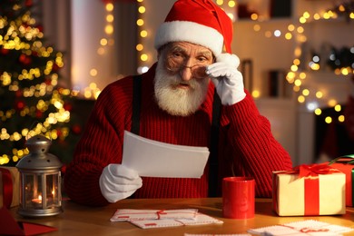 Santa Claus reading letter at his workplace in room with Christmas tree