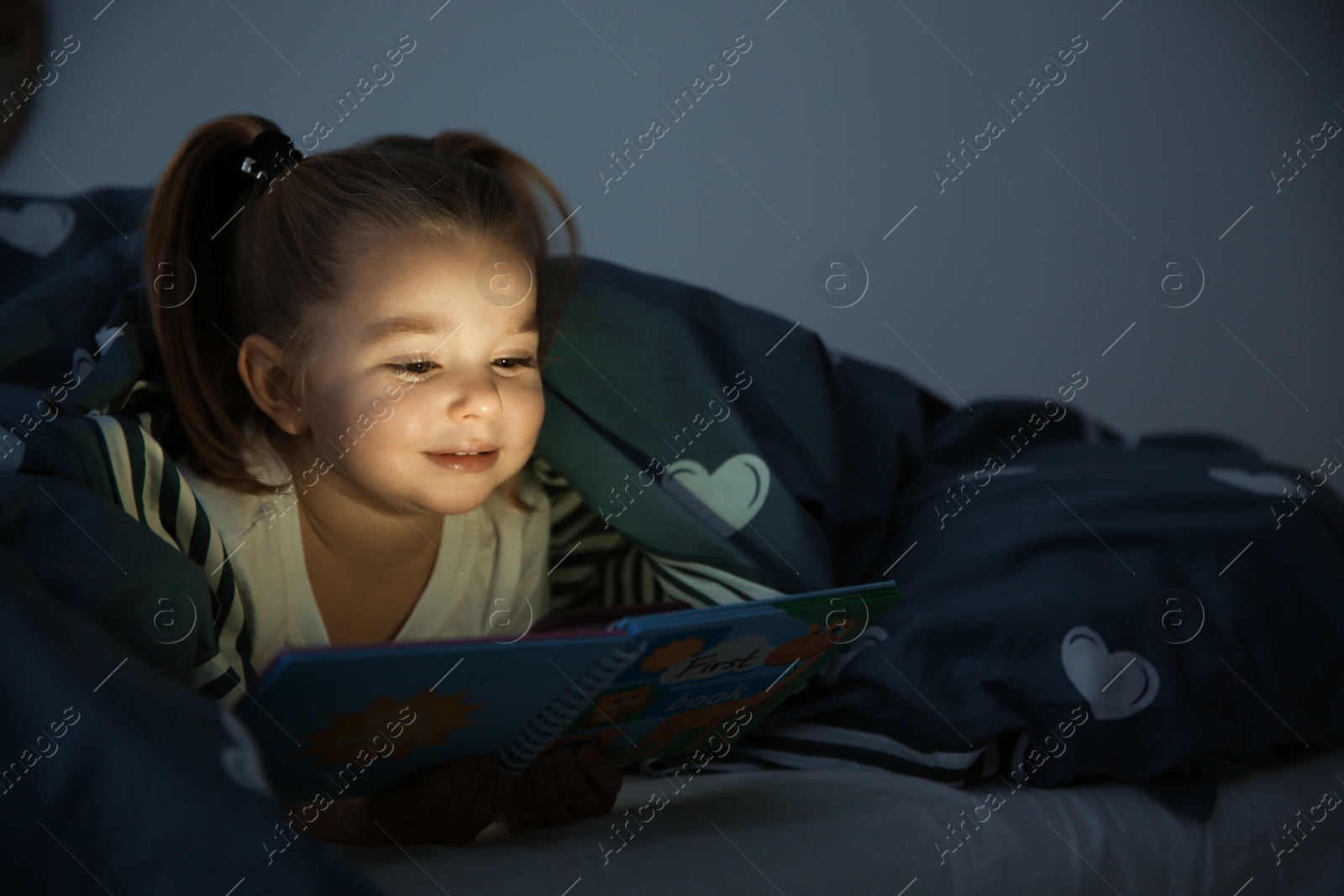 Photo of Beautiful little girl reading book in bed at night. Bedtime story