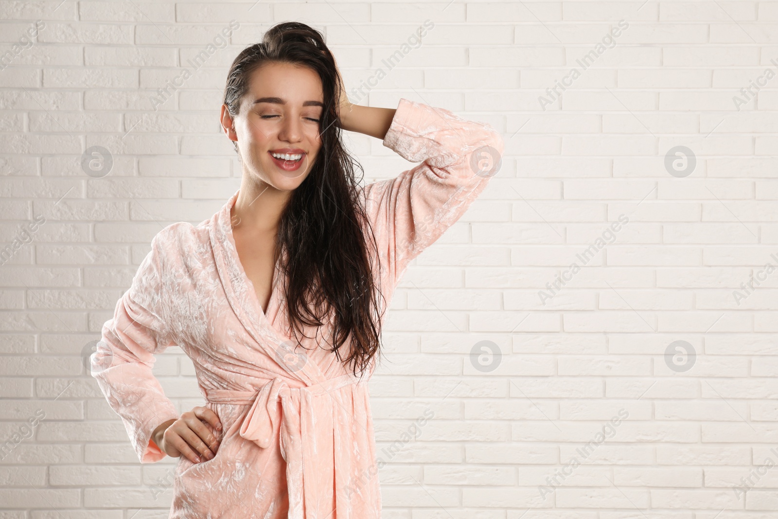 Photo of Young woman in bathrobe with wet hair near white brick wall. Space for text