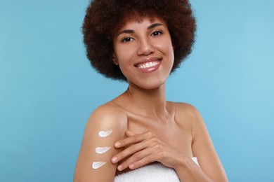 Beautiful young woman applying body cream onto arm on light blue background