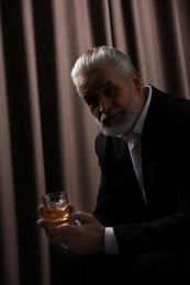 Senior man in formal suit holding glass of whiskey with ice cubes on brown background