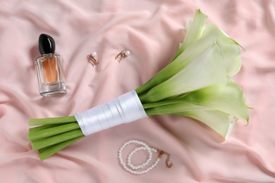 Photo of Beautiful calla lily flowers tied with ribbon, bottle of perfume and jewelry on pink fabric, flat lay