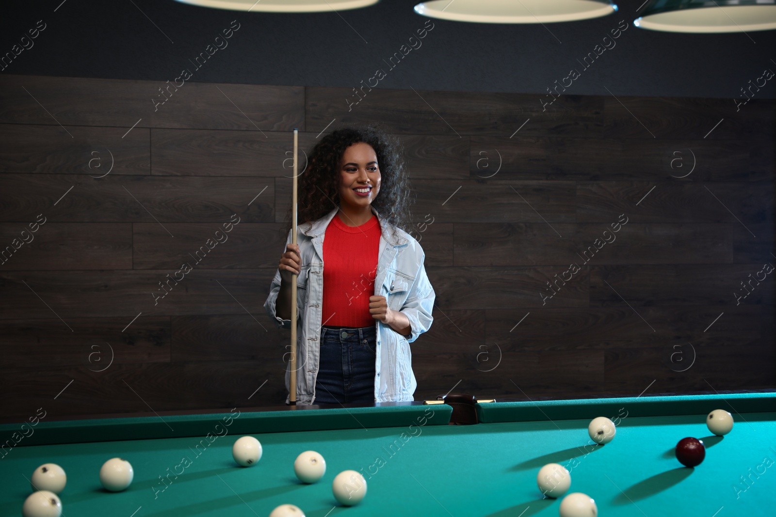 Photo of Young African-American woman with cue near billiard table indoors