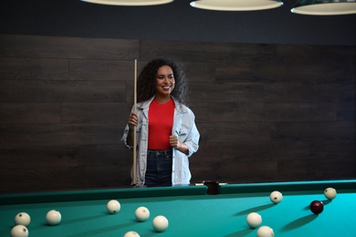 Young African-American woman with cue near billiard table indoors