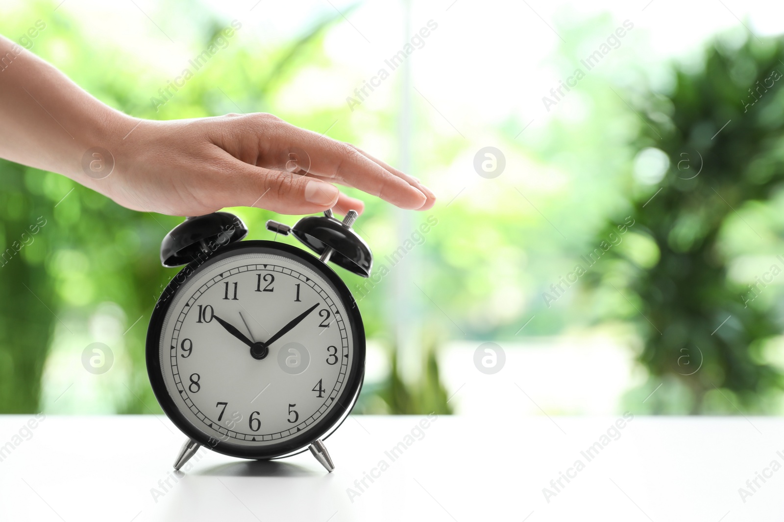 Photo of Woman turning off black alarm clock at white table against blurred background, closeup. Space for text