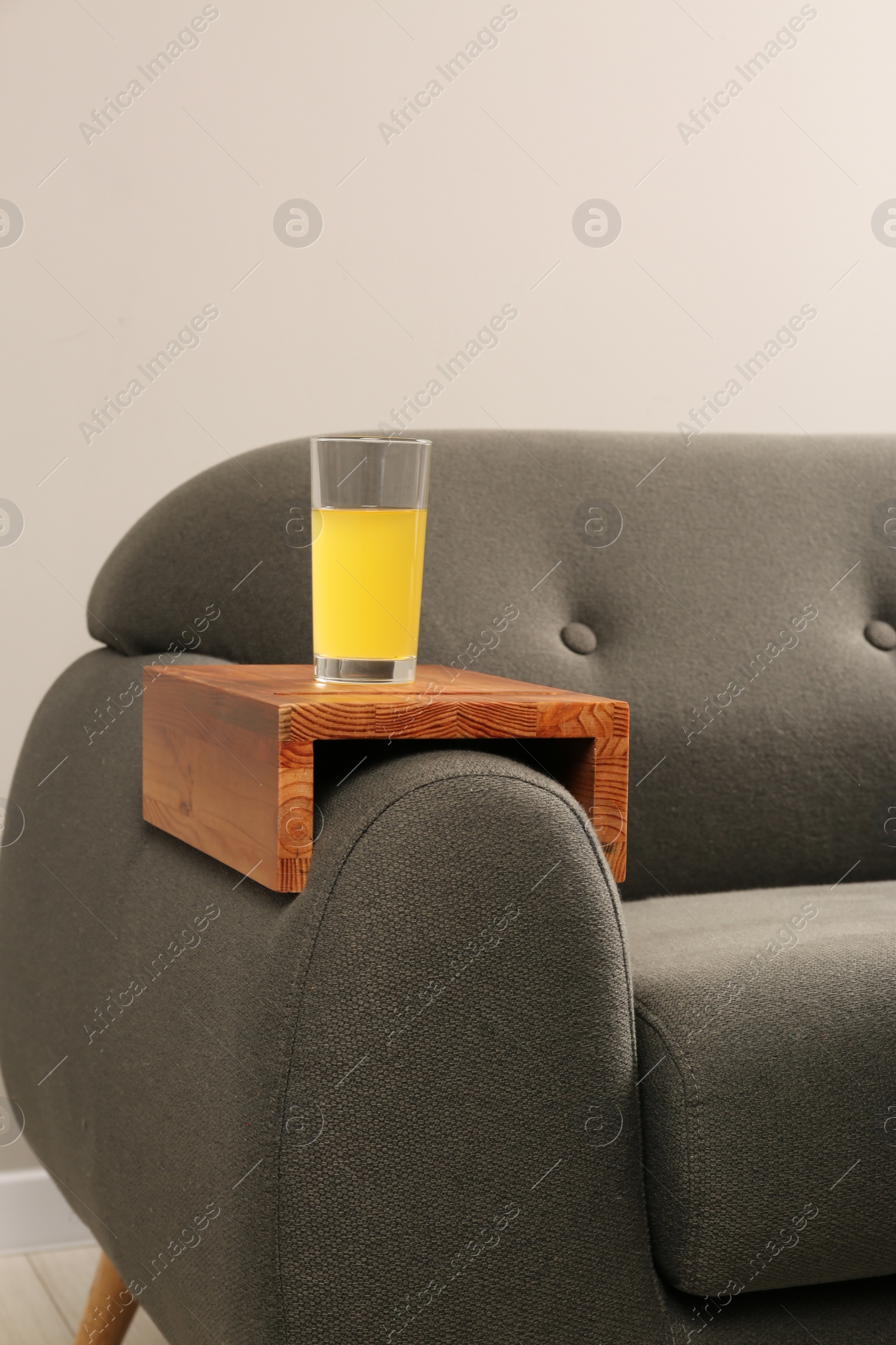 Photo of Glass of juice on sofa with wooden armrest table in room. Interior element