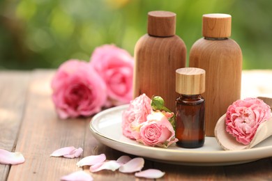 Photo of Bottles of rose essential oil and flowers on wooden table outdoors, space for text