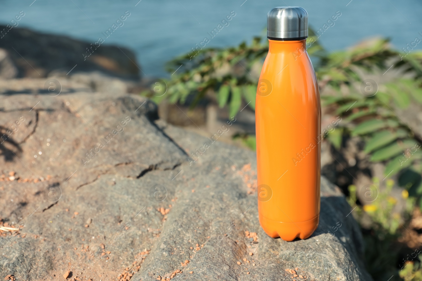 Photo of Modern orange thermos on stone near river. Space for text