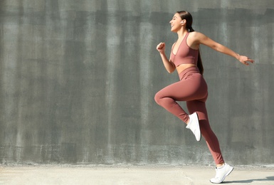 Photo of Happy young woman in stylish sports wear jumping near grey wall. Space for text