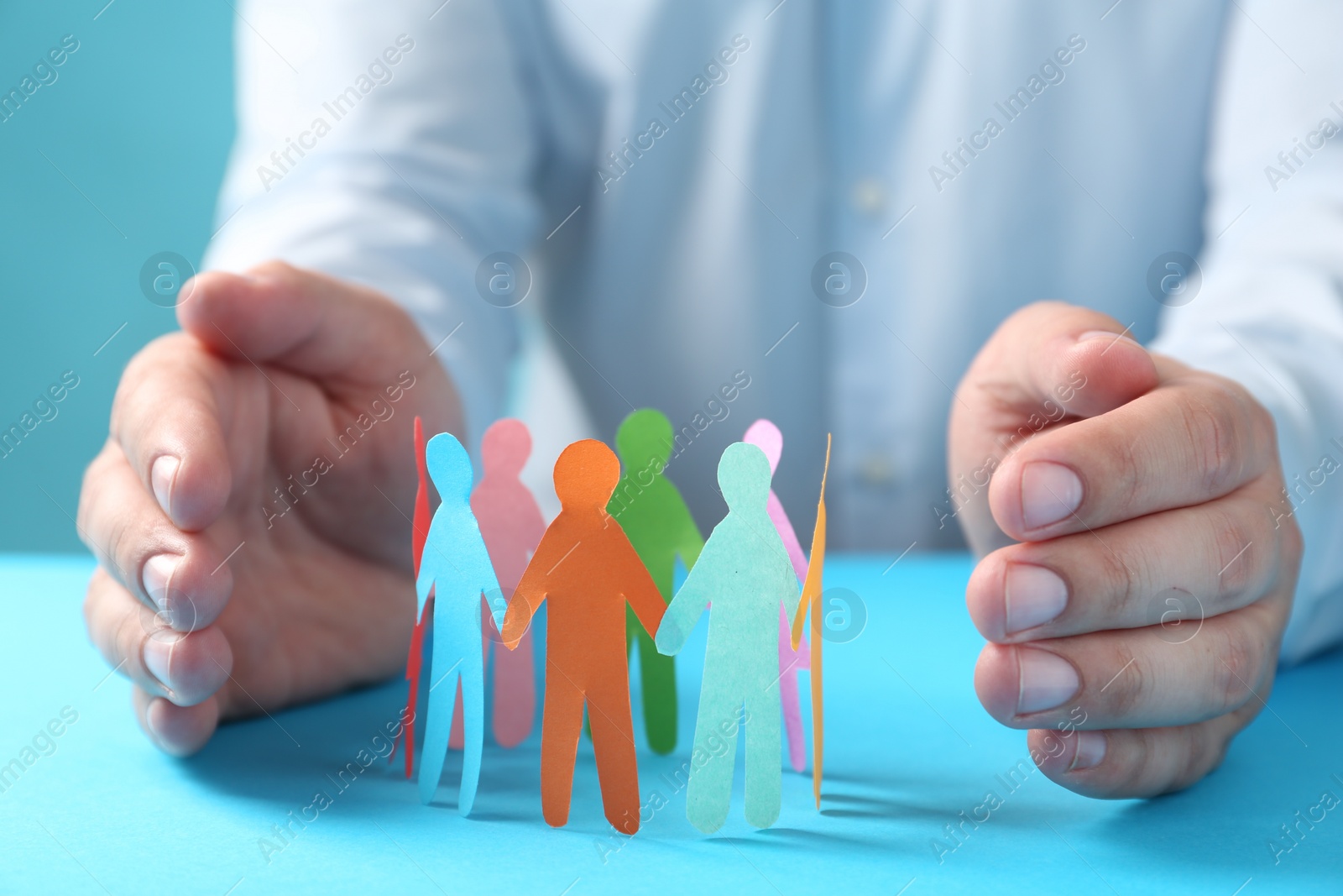 Photo of Man protecting paper human figures on light blue table, closeup. Diversity and inclusion concept