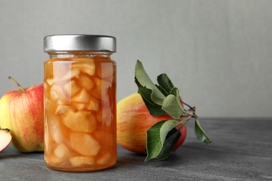 Photo of Tasty apple jam in glass jar and fresh fruits on grey table, space for text