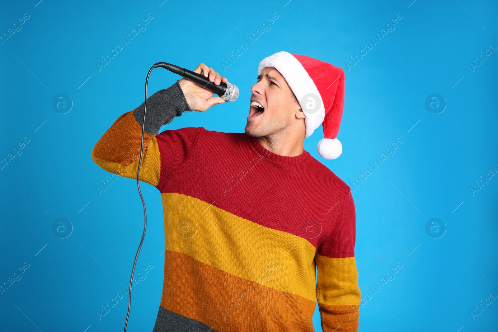 Photo of Emotional man in Santa Claus hat singing with microphone on blue background. Christmas music