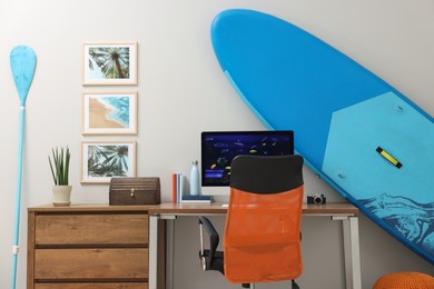 Stylish workplace with modern computer and SUP board near light wall in room. Interior design