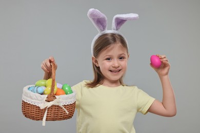 Photo of Easter celebration. Cute girl with bunny ears holding basket of painted eggs on gray background