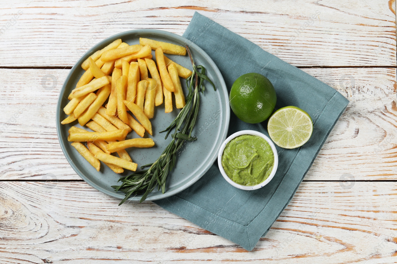 Photo of Plate with delicious french fries, avocado dip, lime and rosemary served on white wooden table, top view