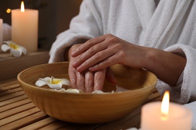 Photo of Woman soaking her hands in bowl of water and flowers at wooden table, closeup. Spa treatment