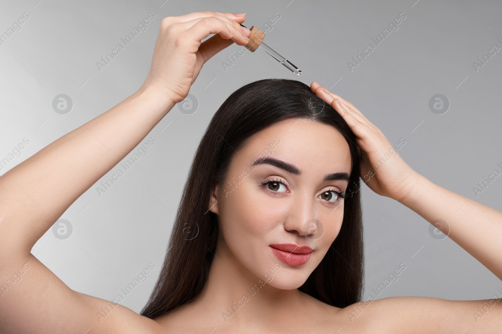 Photo of Young woman applying essential oil onto hair roots on light grey background