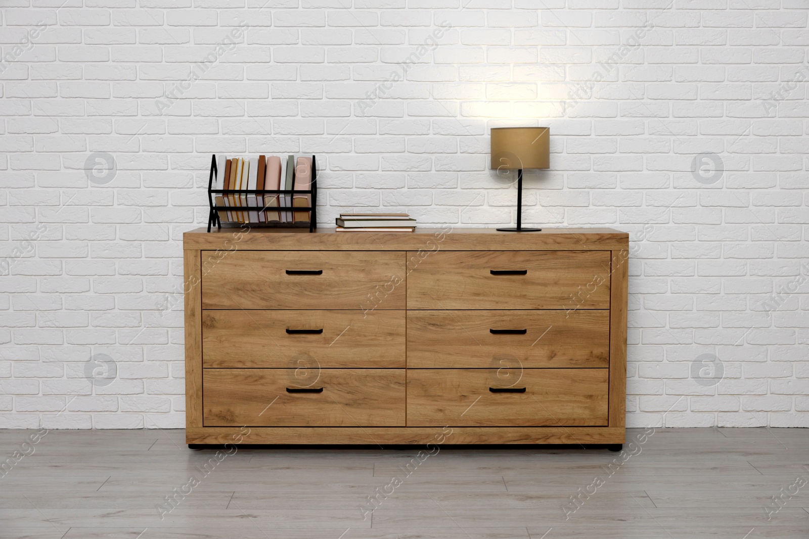 Photo of Wooden chest of drawers, books and lamp near white brick wall. Interior design