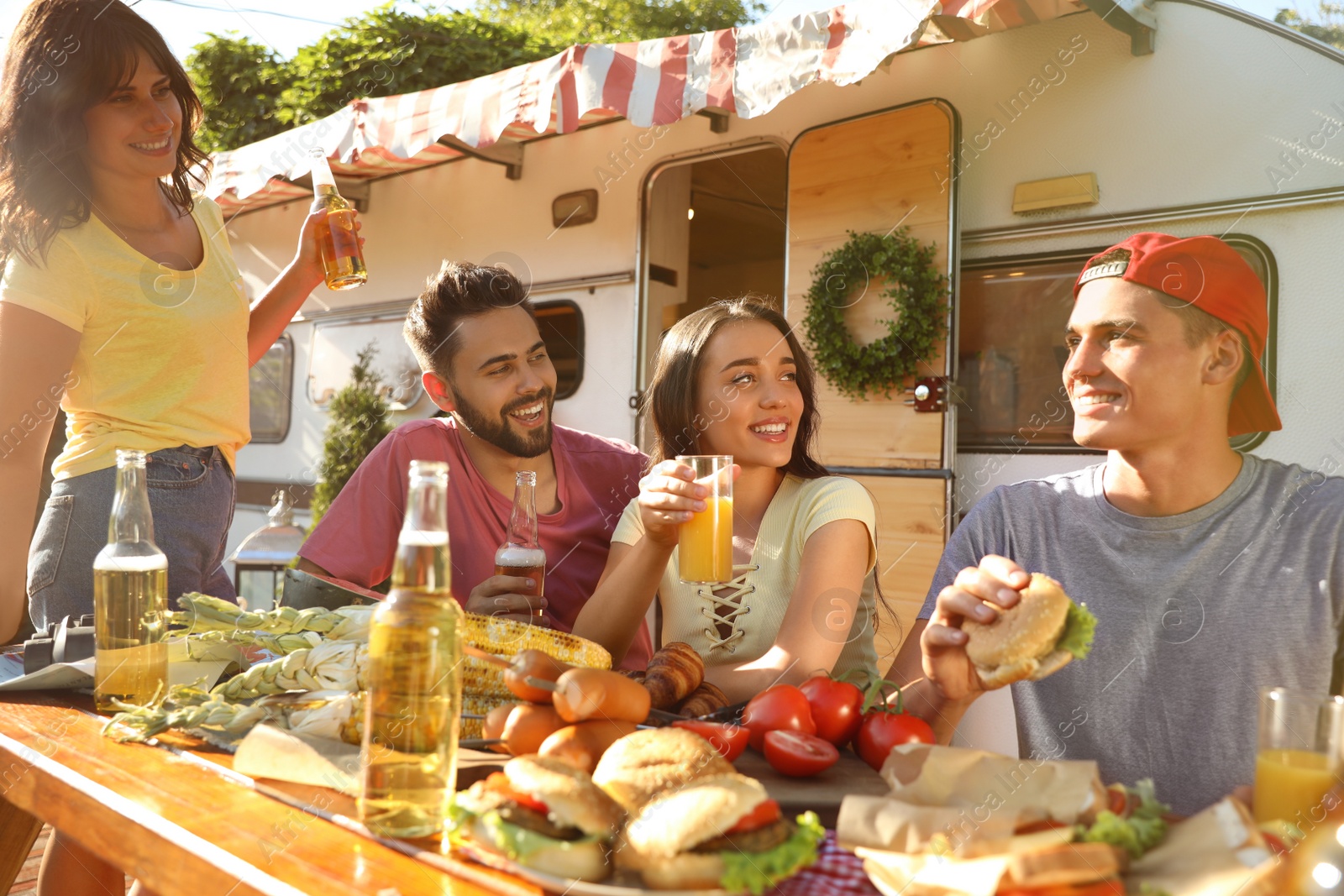 Photo of Happy friends with drinks and food at table near motorhome. Camping season