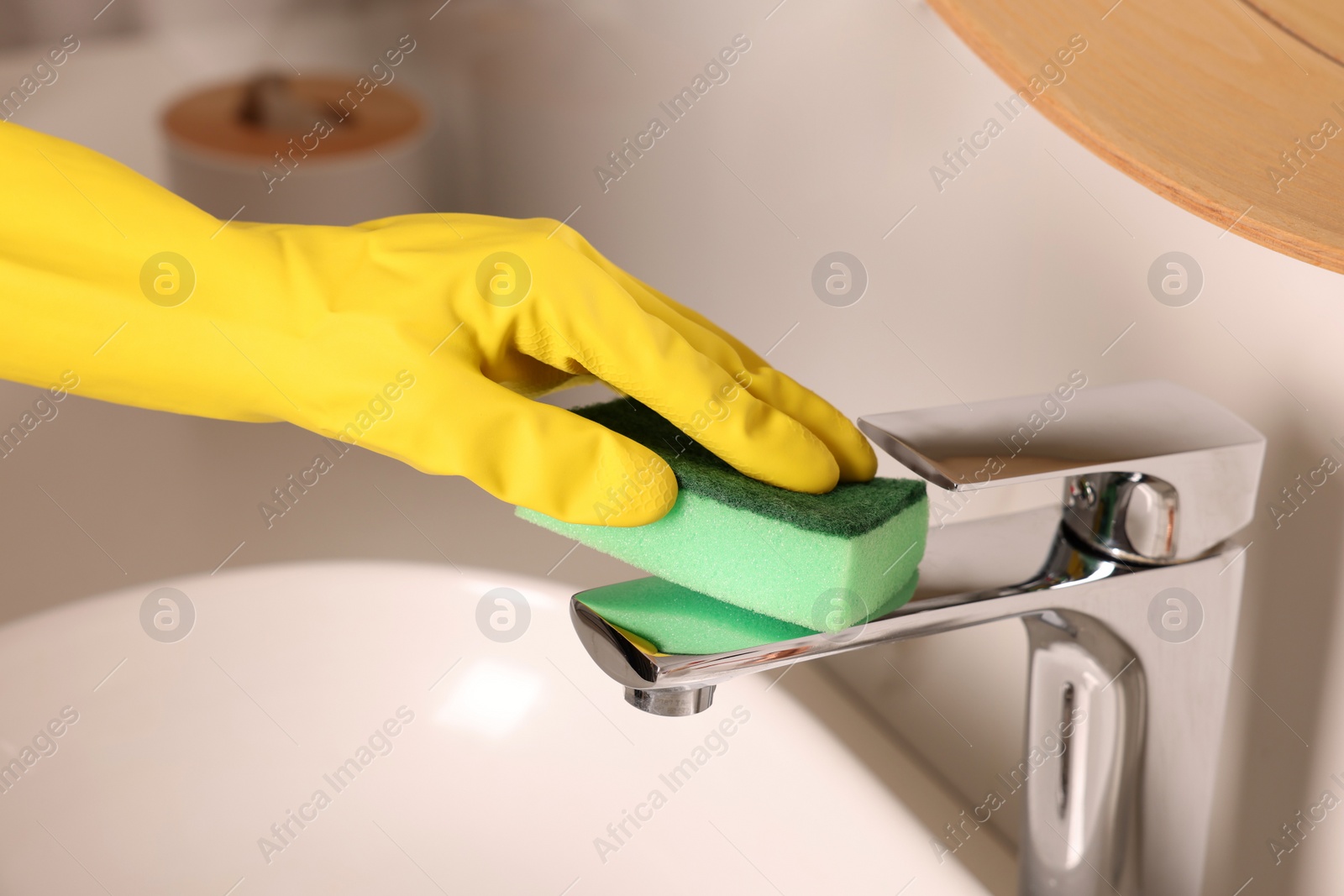 Photo of Woman in gloves cleaning faucet of bathroom sink with sponge, closeup