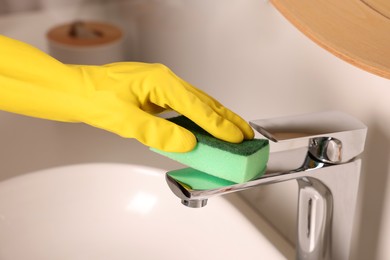 Photo of Woman in gloves cleaning faucet of bathroom sink with sponge, closeup