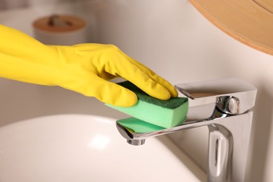 Woman in gloves cleaning faucet of bathroom sink with sponge, closeup