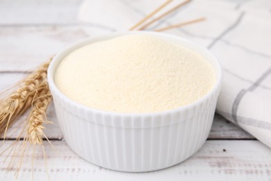 Photo of Uncooked organic semolina in bowl and spikelets on white wooden table, closeup