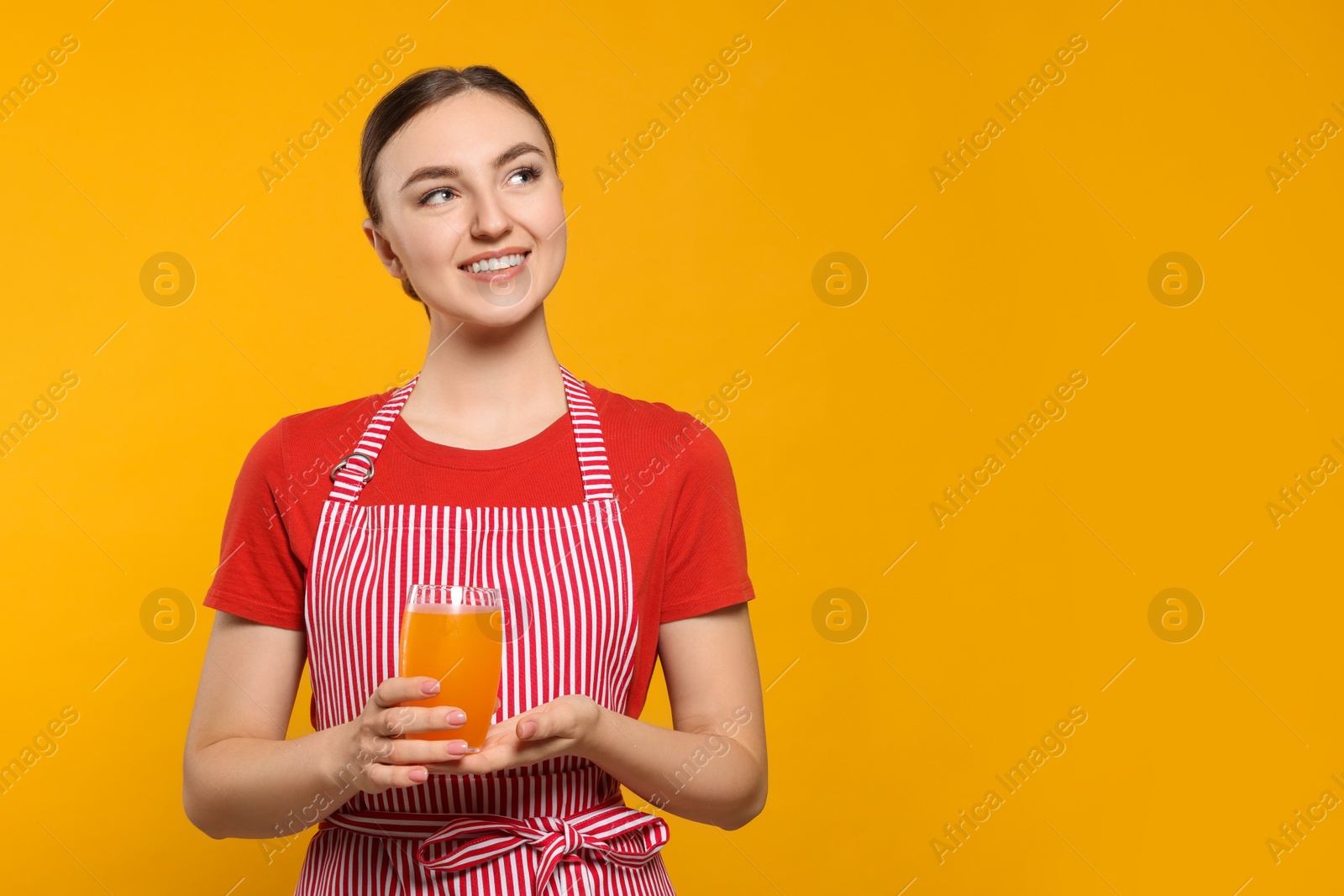 Photo of Beautiful young woman in clean striped apron with glass of juice on orange background. Space for text