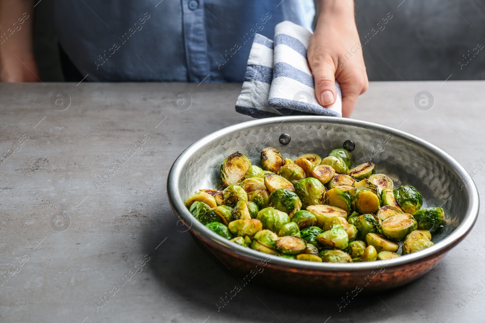 Photo of Woman with frying pan of roasted Brussels sprouts at grey table, closeup