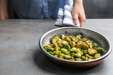 Photo of Woman with frying pan of roasted Brussels sprouts at grey table, closeup