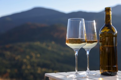 Photo of Glasses and bottle of white wine on wooden table against mountain landscape, space for text