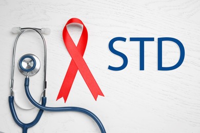 Image of Red awareness ribbon, stethoscope and abbreviation STD on white wooden background, flat lay