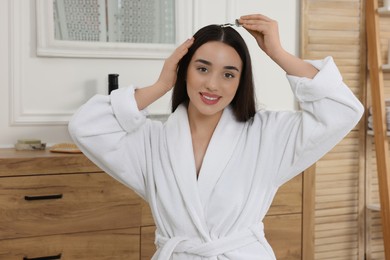 Photo of Happy young woman applying essential oil onto hair roots in bathroom