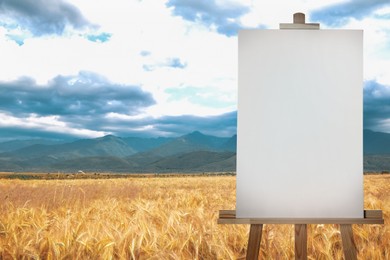 Wooden easel with blank canvas in wheat field on cloudy sky. Space for text