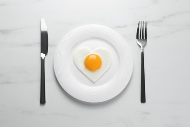 Plate with heart shaped fried egg and cutlery on white marble table, flat lay