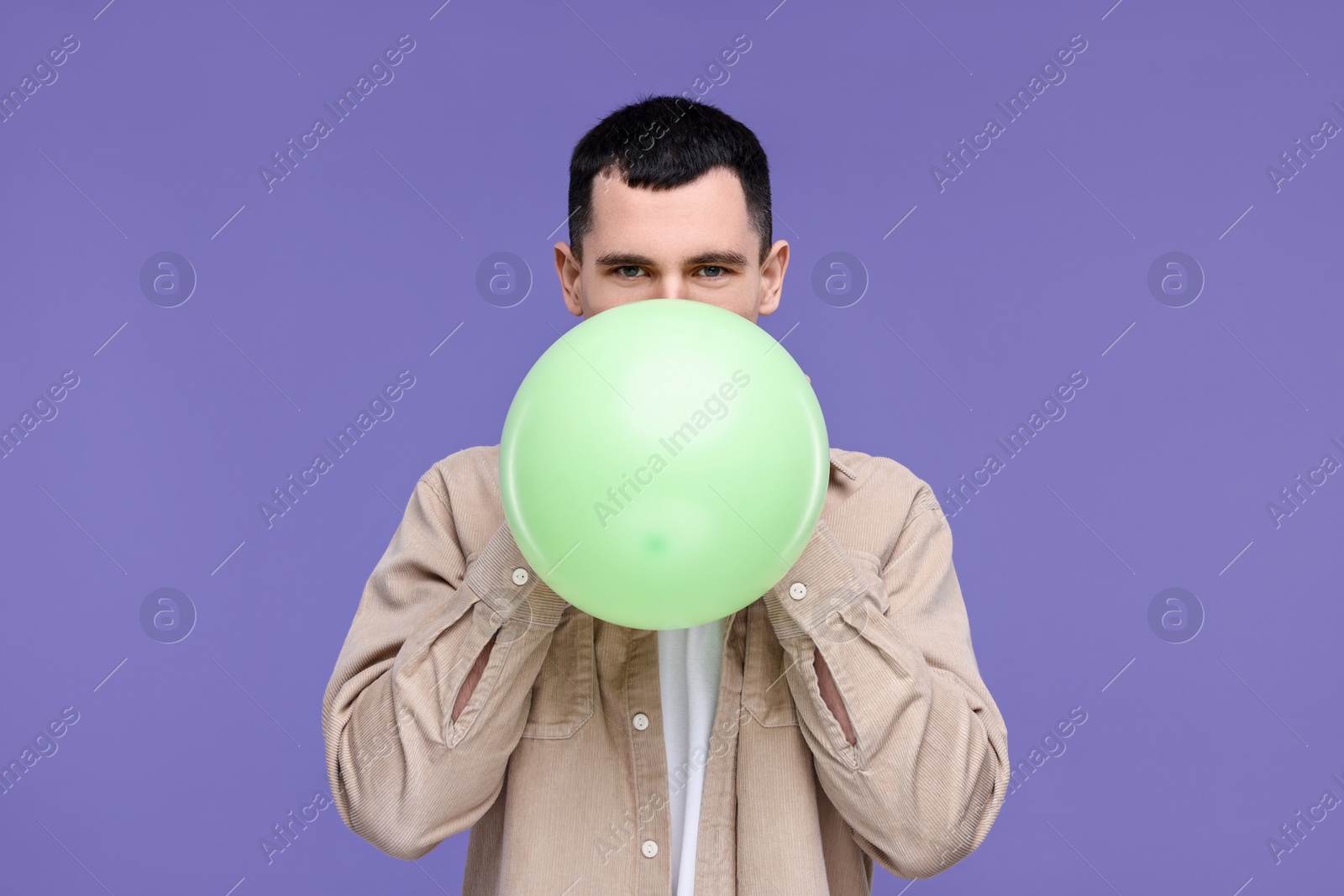 Photo of Young man inflating light green balloon on purple background