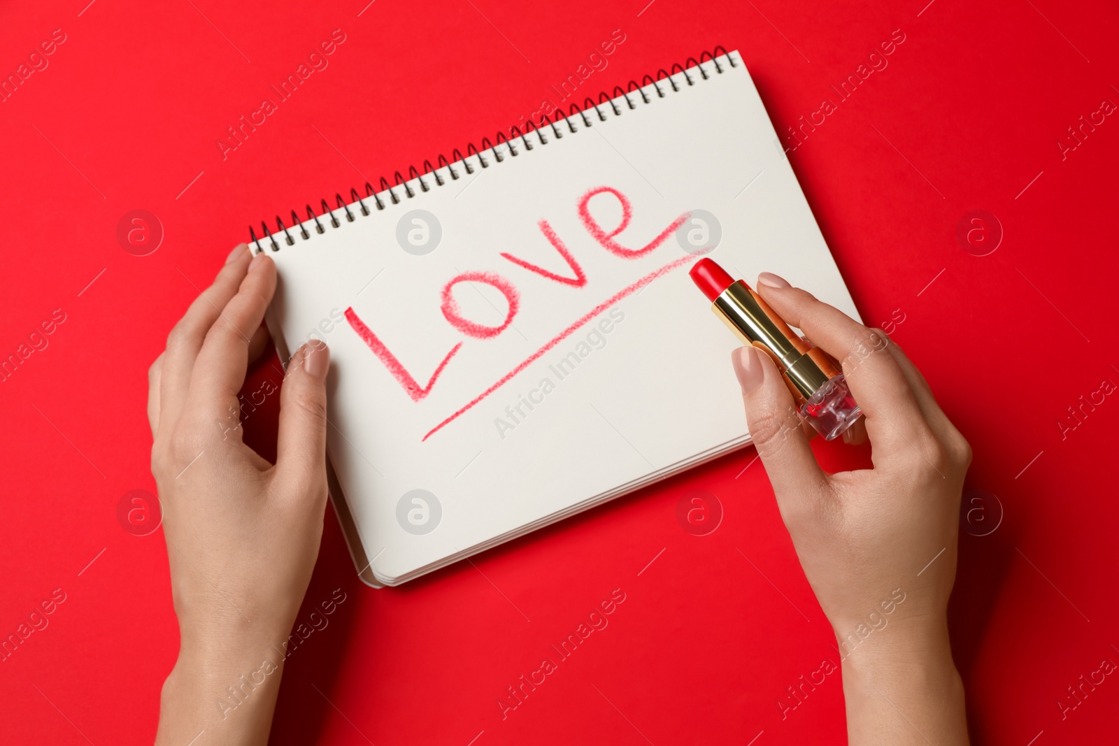 Photo of Woman writing word LOVE with lipstick in notepad on red background, top view