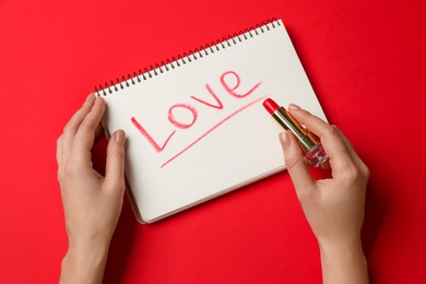 Photo of Woman writing word LOVE with lipstick in notepad on red background, top view