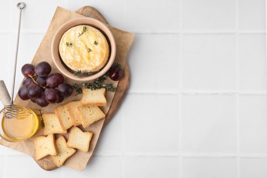 Photo of Tasty baked camembert in bowl, croutons, grapes, honey and thyme on white tiled table, top view. Space for text