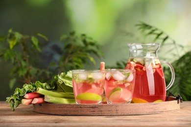 Photo of Glasses and jug of tasty rhubarb cocktail with citrus fruits on wooden table outdoors