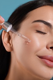 Photo of Beautiful young woman applying serum onto her face on light blue background, closeup